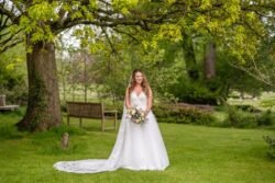 A bride in a white dress holding a bouquet, smiling in the lush gardens of Orchardleigh House with trees and a bench in the background.