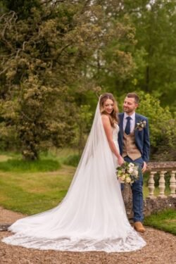 Bride in a long white veil and groom in a blue suit smiling and holding hands at an Orchardleigh House garden wedding.