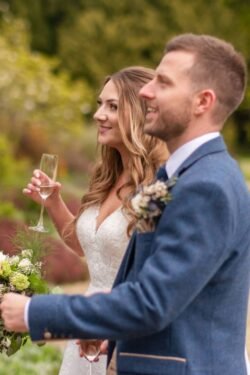 A bride in a white dress and a groom in a blue suit holding champagne glasses at an Orchardleigh House outdoor wedding ceremony.