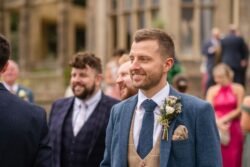At an Orchardleigh House wedding, a man in a blue tweed suit with a boutonnière smiles, surrounded by guests in formal attire.