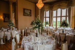 Elegant dining room setup for an Orchardleigh House Wedding, featuring floral centerpieces on white clothed tables, ornate curtains, and a chandelier in a room with large windows and classic paintings.