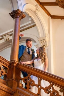 A bride and groom smiling at each other on an ornate wooden staircase in Orchardleigh House, with the groom reaching towards the bride.