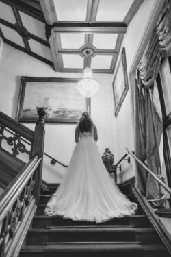 A bride in a long white gown ascends a grand staircase at Orchardleigh House, highlighted by a large chandelier above.