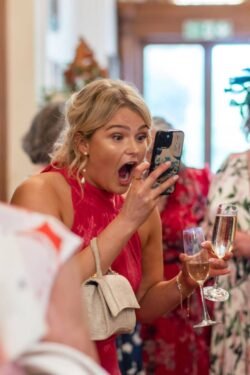 A surprised woman in a red dress holding a champagne flute and looking at a smartphone at an Orchardleigh House Wedding.
