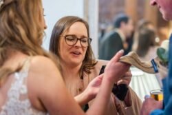 A woman in glasses and a beige jacket engages in conversation with other guests at an Orchardleigh House Wedding, gesturing towards a smartphone being held by another person.