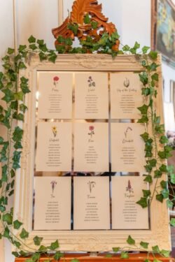 Elegant seating chart displayed in a vintage gold frame, adorned with green vines, featuring floral illustrations and names for Orchardleigh House Weddings.