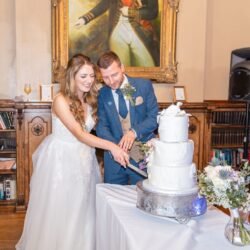 Bride and groom smiling as they cut a multi-tiered white wedding cake together at Orchardleigh House, surrounded by classical decor.