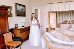A bride in a white dress smiling in an elegant bedroom at Orchardleigh House, with a canopy bed and antique furniture.