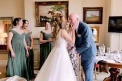 A bride whispers to her father at Orchardleigh House, surrounded by bridesmaids in green dresses in an elegant room, with emotional expressions on their faces.