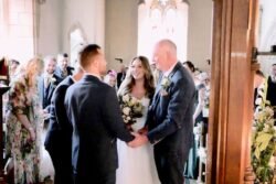 Bride and groom holding hands at Orchardleigh House, exchanging vows as guests watch in a church with ornate architecture.