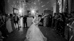 A couple performs a dance dip in the center of a ballroom at Orchardleigh Weddings, surrounded by guests who are watching and taking photos. The room is dimly lit with decorative lights on the walls, creating a romantic ambiance.