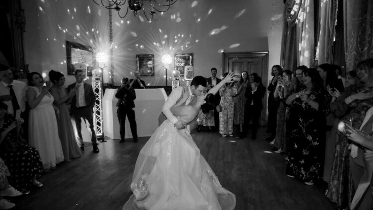 A couple performs a dance dip in the center of a ballroom at Orchardleigh Weddings, surrounded by guests who are watching and taking photos. The room is dimly lit with decorative lights on the walls, creating a romantic ambiance.