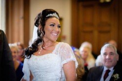 A bride in a white dress and tiara smiles during an Orchardleigh Weddings ceremony, with guests seated in the background.