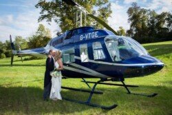 A bride and groom kiss beside a blue helicopter with registration number G-VTGE in a grassy field at Orchardleigh Weddings. Trees and a partly cloudy sky form the perfect backdrop.