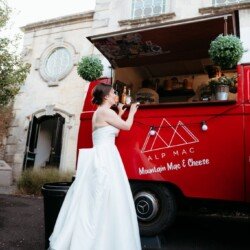 A woman in a white wedding dress stands by a red food truck labeled "ALP MAC Mountain Mac & Cheese" near the stone building of Rook Lane Weddings Frome.
