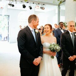 At a Rook Lane Weddings Frome ceremony, a bride, holding a bouquet, stands between two men in formal attire, with guests seated and standing in the background.