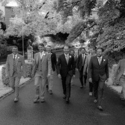 A group of seven men in suits, some with boutonnieres, walk down a tree-lined path. They are smiling and appear to be part of a wedding party at Rook Lane Weddings Frome.