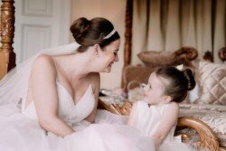 A bride in a white dress smiles at a young girl wearing a matching outfit as they sit on an ornately carved wooden bench, capturing the timeless elegance that Orchardleigh House Weddings are known for.