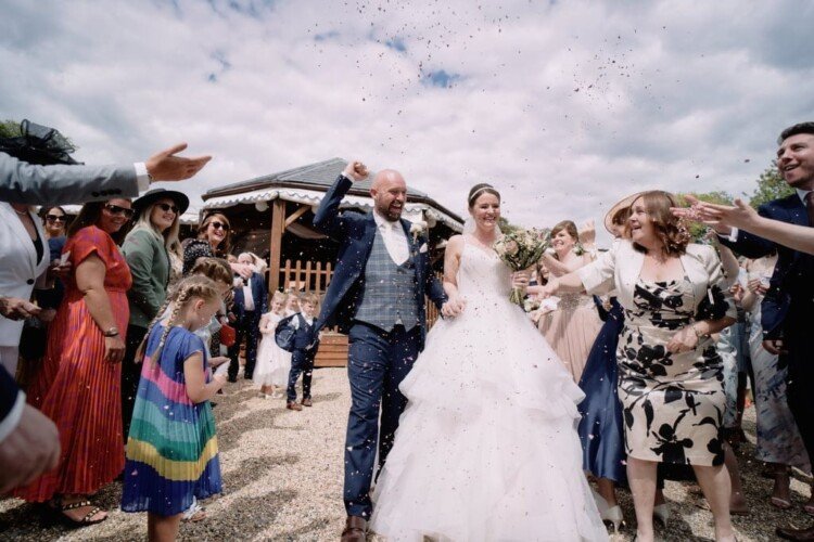 A newlywed couple walks down a path at Orchardleigh House Weddings, surrounded by guests throwing confetti. The bride is in a white gown and the groom in a blue suit. Celebrants are smiling and cheering under a partly cloudy sky.