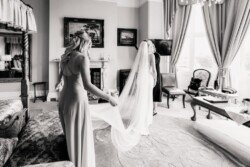 A bride in a long veil stands in the beautifully decorated room of Orchardleigh House Weddings, while another woman, possibly a bridesmaid, adjusts her dress.
