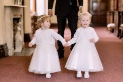 Two young girls wearing white dresses and flower crowns hold hands while walking down a carpeted aisle at an Orchardleigh House wedding, with an adult in the background.