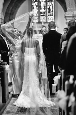 www.thefxworks.co.uk A bride and groom stand at the altar in a church. A woman adjusts the bride's long veil. The scene, reminiscent of an Orchardleigh House Wedding, is in black and white.