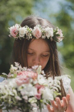 A young girl with long hair wears a flower crown and a white dress, looking down at a bouquet of flowers. Green foliage is blurred in the background, capturing the essence of Orchardleigh House Weddings.