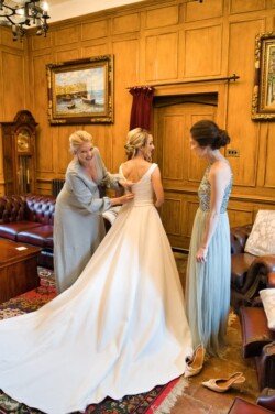 Three women are in a wood-paneled room. One is wearing a wedding dress being adjusted by another woman in a robe, while the third woman stands nearby in a formal dress, capturing the timeless elegance typical of Orchardleigh House Weddings.