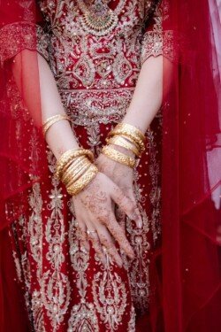 A person wearing an ornate red and gold traditional outfit with intricate henna designs on their hands and multiple gold bangles could be seen at the picturesque Orchardleigh House Weddings.