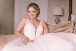 A woman in a white wedding dress sits on a sofa, smiling at the camera. A lamp and a wooden bedpost are visible in the background, reflecting the elegance of Orchardleigh House Weddings.