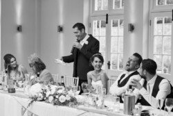 www.thefxworks.co.uk A man speaks into a microphone, smiling, at an Orchardleigh House Weddings reception. The bride and other guests at the long table also smile and laugh. The table is adorned with flowers, drinks, and glasses.