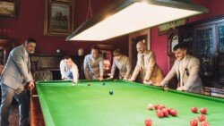 Six men in suits are gathered around a billiard table in a dimly lit room, appearing engaged in a game. The room, reminiscent of Orchardleigh House Weddings, is adorned with framed paintings and antique furniture, adding to its classic charm.