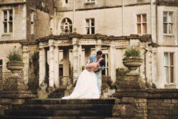 A couple dressed in wedding attire poses on stone steps, with the groom dipping the bride in front of the grand Orchardleigh House, its old stone building adorned with columns and ornate details.