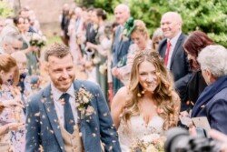 A bride and groom walk down an outdoor aisle filled with guests who are throwing flower petals at their Orchardleigh House wedding. The groom wears a blue suit, and the bride wears a white dress, both smiling.