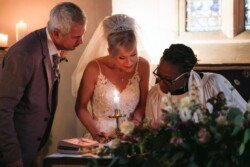 Three people, including a bride in a white dress and veil, are signing papers near a candle in the dimly lit church setting adorned with flowers. The intimate moment at Orchardleigh House Weddings captures the beauty and solemnity of the occasion.