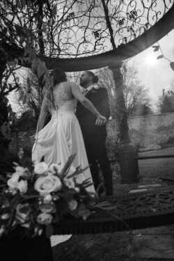 A bride and groom stand holding hands under an ornate outdoor structure at Orchardleigh House Weddings, with the bride in a flowing dress and the groom in a suit. A bouquet of white roses is in the foreground.