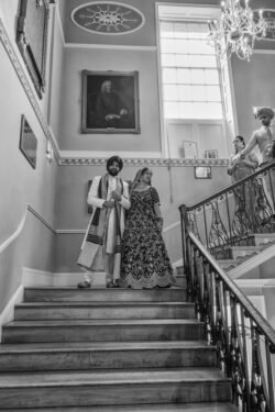 A bride and groom in traditional attire descend a staircase inside the grand Guildhall, with three onlookers standing at the railing above. Bath Wedding Photographer captures the timeless moment perfectly.