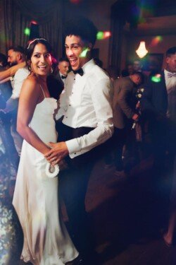 A couple dancing at a formal event at Orchardleigh House Weddings, dressed in a white wedding gown and a white shirt with black pants, surrounded by other guests in a dimly lit room with colorful lights.