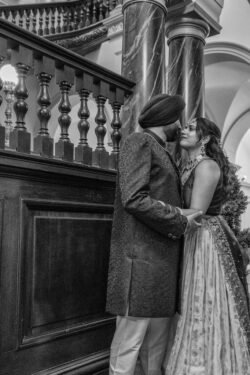 Black and white photo of a couple in formal attire standing close to each other by a staircase at the Guildhall Bath, holding hands and gazing at each other with a slight smile.