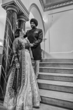 A couple in traditional attire stand on a staircase; the woman in a lehenga and the man in a sherwani and turban, looking at each other affectionately. Captured by Guildhall Bath Wedding Photographer, this black and white photograph exudes timeless romance.