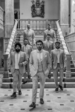 A group of eight men in suits walk confidently down a staircase in a formal setting at Farleigh House, with one man leading at the front. Black and white photograph captured by a wedding photographer.