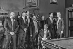 A group of seven men dressed in formal suits pose in a room at Orchardleigh House with dark walls and classic decor, including framed paintings and a billiard table in the foreground.