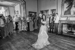 A couple shares their first dance in a large, elegantly decorated room at Orchardleigh House while guests stand around watching and clapping. The setting features ornate frames and a fireplace. Black and white photograph.