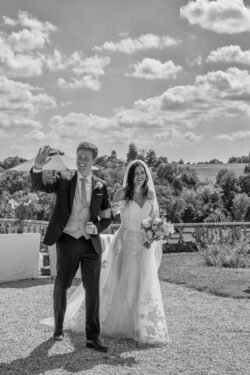 A bride and groom walk outdoors in wedding attire at Orchardleigh House; the groom waves while holding a drink, and the bride carries a bouquet. The background includes a scenic landscape with trees and clouds.