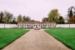 A symmetrical brick building with large windows sits elegantly in a manicured garden, perfect for an Elmhay Park wedding photographer. A gravel path leads to the entrance, flanked by lush green lawns and trees in the background.