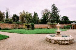 A walled garden with a circular stone fountain and gravel paths sets the scene. Lush grass, trees, and a wooden gate add charm under an overcast sky. Imagine capturing an Elmhay Park wedding here, where each moment is framed by nature’s serene beauty.