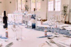 The dining table, perfect for an Elmhay Park wedding photographer's capture, boasts wine bottles, elegant glassware, and a centerpiece featuring blue accents and a white rose, all beautifully illuminated by the sunlight streaming through the large windows.