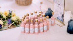 Small pink bottles with gold caps are elegantly arranged on a table, surrounded by floral decorations and a framed sign. Capturing every detail, the Elmhay Park wedding photographer ensures these charming touches are remembered forever.