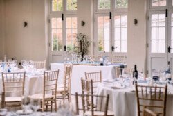 A wedding reception setup at Elmhay Park with round tables and chairs, white tablecloths, wine bottles, glasses, and a centerpiece featuring "Mr & Mrs" signage. Sunlight illuminates the room through large windows.