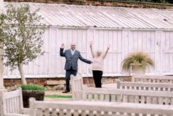 Two people, undoubtedly celebrating an Elmhay Park Wedding, stand outside smiling and waving in front of a white greenhouse. A charming wooden fence and potted plants add to the foreground's quaint allure.